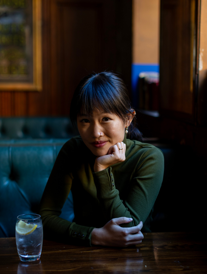 Woman in green top sits at a table with a glass of water and lemon, looking at the camera.