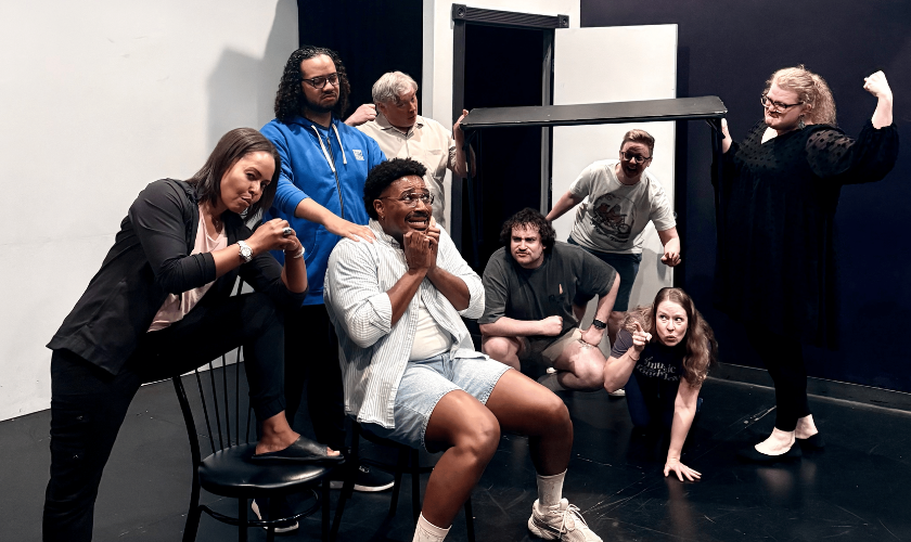 Eight people pose dramatically in a theater with chairs and a door frame on a black stage.