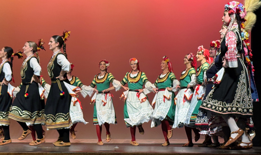 Dancers in folk costumes perform on stage against an orange background.