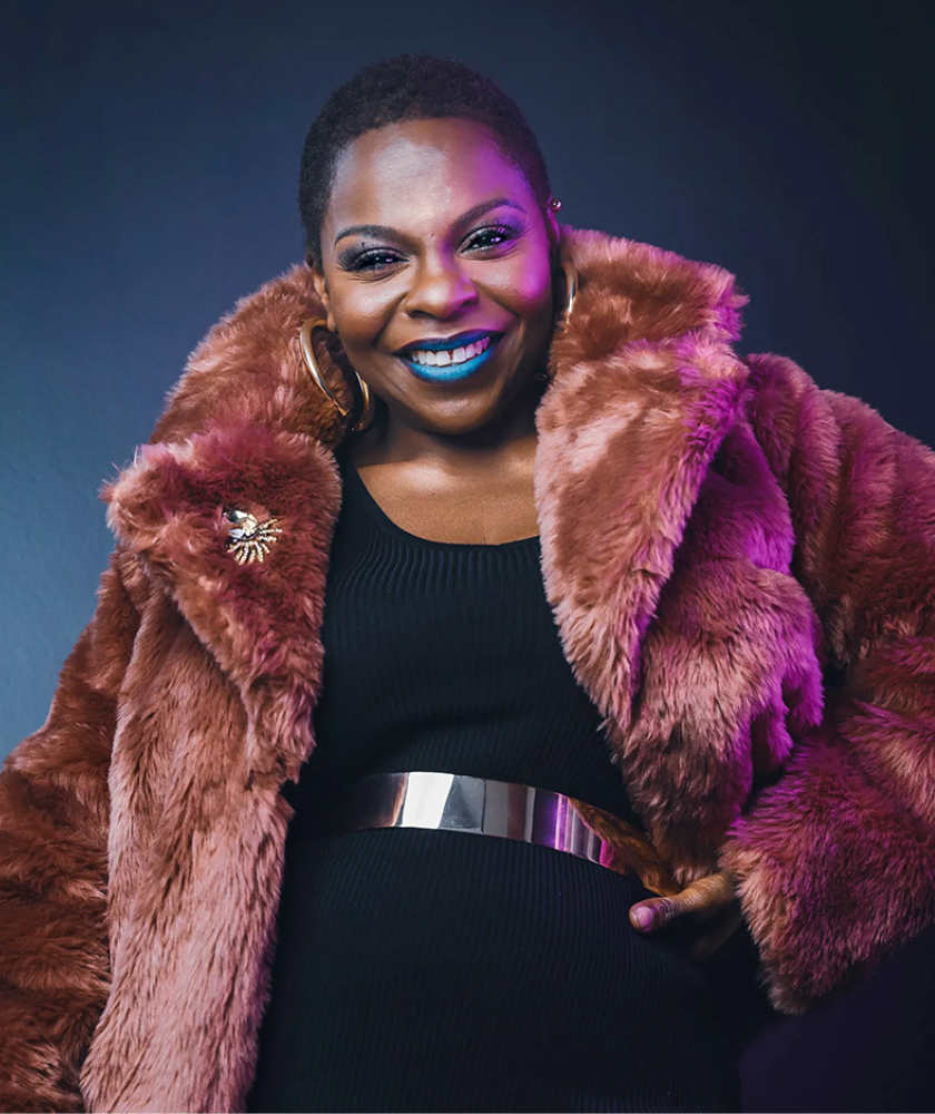 Smiling woman in a faux fur coat, black dress, and bold blue lipstick against a dark background.