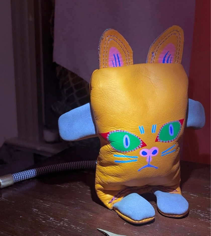 A plush toy with a cat face, yellow body, blue arms, and colorful eyes sits on a table.