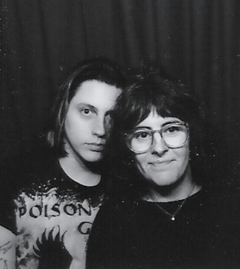 Two people pose in front of a dark curtain, one wearing glasses and both looking at the camera.