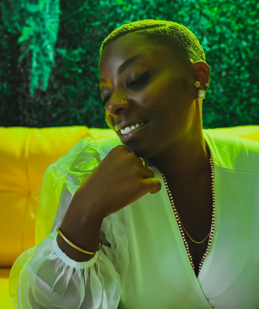 A woman in white with dark skin and light hair smiles softly with eyes closed, sitting on a yellow couch with a green background.