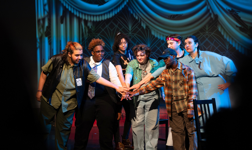 A group of six people on stage, standing close together with their hands stacked in the center, as if in a team huddle, under dramatic lighting and a decorative backdrop.