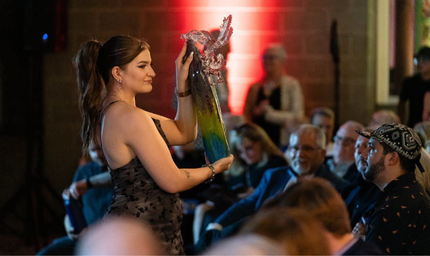 A person in a formal dress holds up a colorful glass sculpture on stage in front of an audience, with red lighting in the background.