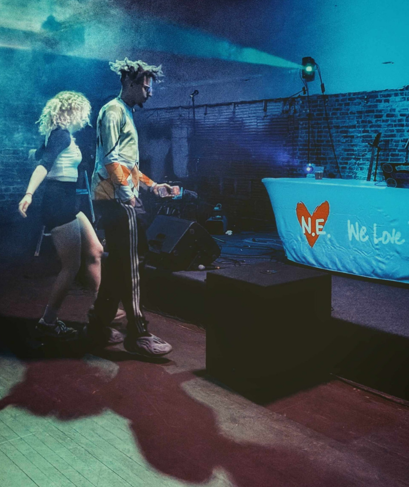 Two people walk through a dimly lit venue with stage lights and smoke. A table with a white cloth and heart logo reading N.E. We Love is in the background against a brick wall.