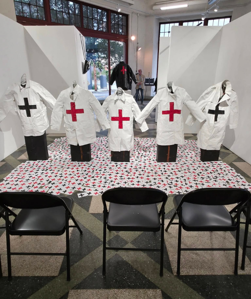 Five white lab coats with black and red crosses are displayed upright in a gallery, arranged in a row on patterned flooring. Three black chairs are set in front, and large windows are visible in the background.