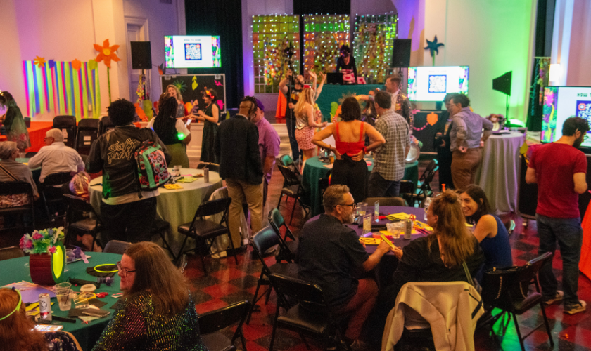 A lively indoor party with people socializing at decorated tables, colorful lights, and vibrant wall decorations. A DJ plays music on a stage in the background while guests mingle, eat, and enjoy the festive atmosphere.