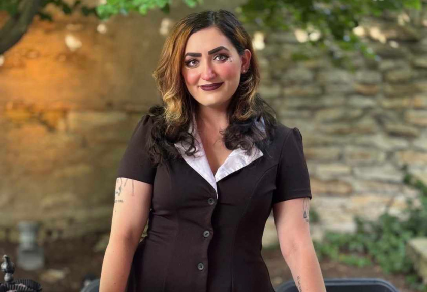 Person with wavy hair in black top in front of a stone wall.