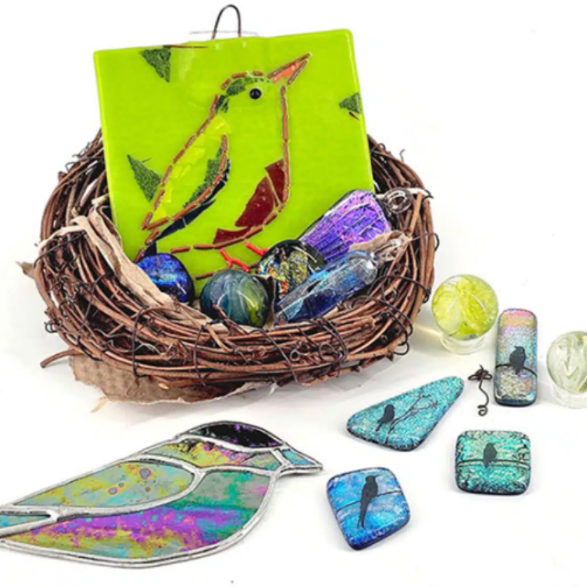 Colorful glass bird art in a twig nest with fused glass jewelry and marbles on a white background.