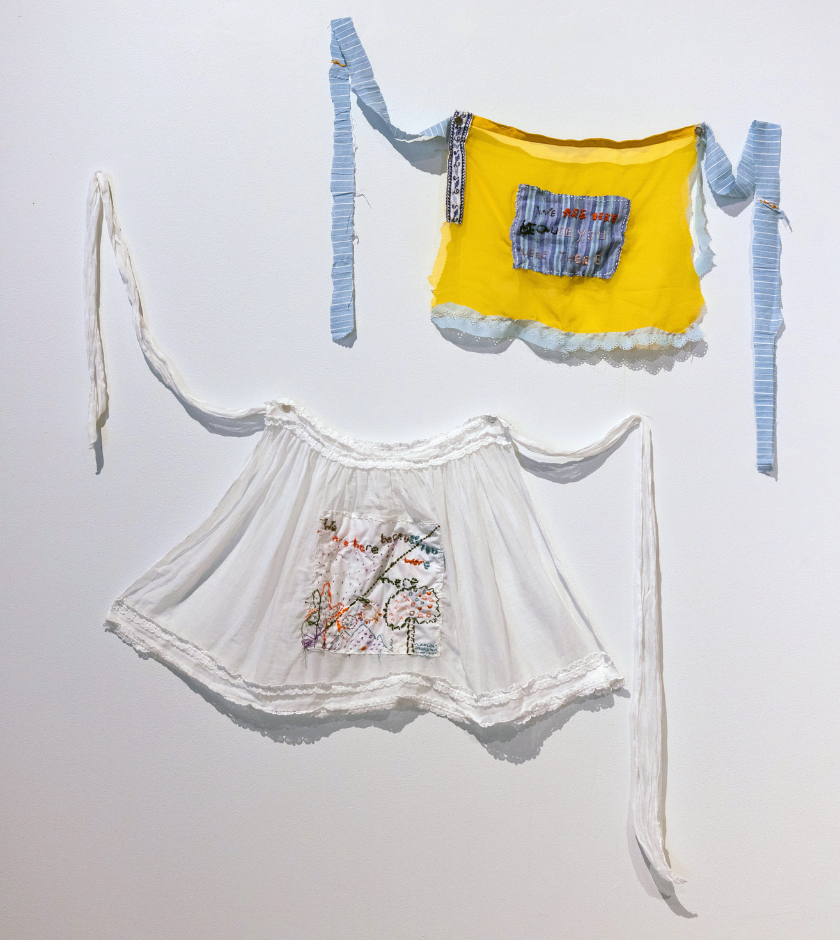 Two fabric aprons, one yellow and one white, with blue and white straps, hang on a wall.