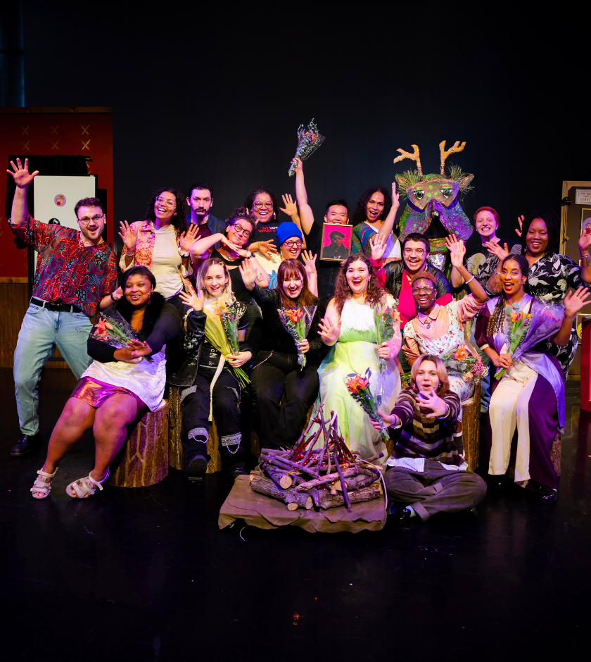 A group of people in colorful costumes pose and smile on a stage with props and flowers.