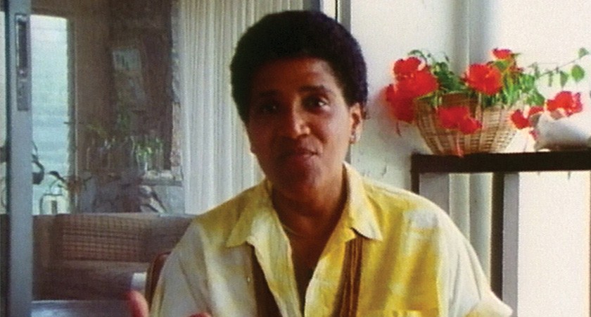 A person with shortly cropped dark hair and a yellow-and-white top sits inside a home with a couch and a basket of flowers on a table visible in the background