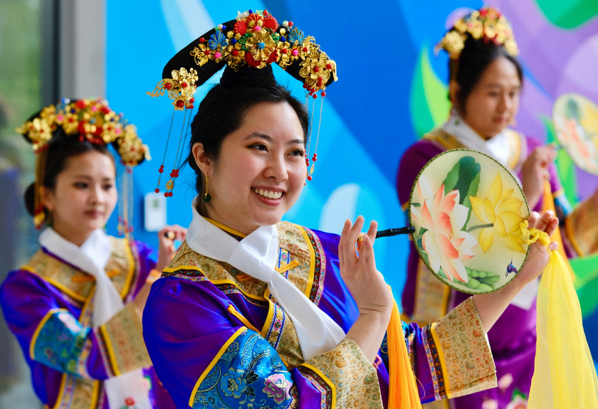 Three women in ornate Chinese costumes perform a traditional dance, one holding a decorated fan.