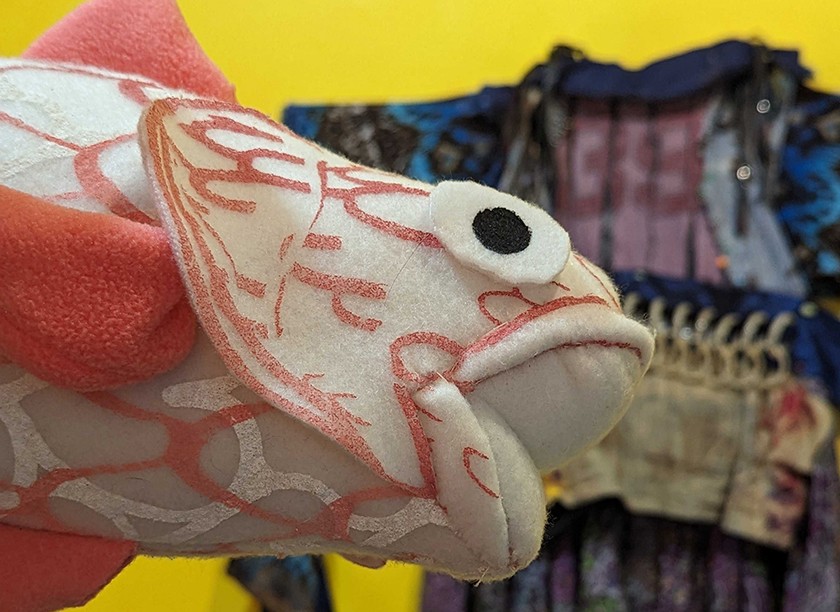 A pink-and-white fish made out of fabric is shown in the foreground.  Blurred in the background is a multi-colored shirt hanging on a bright yellow gallery wall