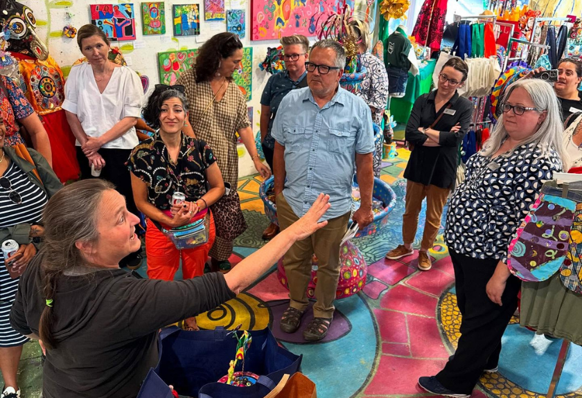A group listens to a woman speaking in a colorful, art-filled room with vibrant bags and art.