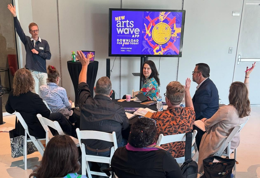People at a workshop, some raising hands, with Arts Wave app display on screen.
