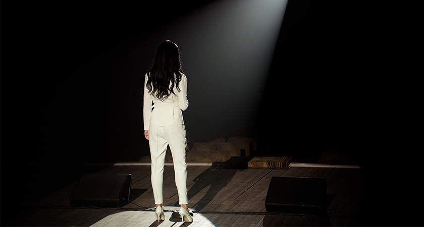 A person with long dark hair, wearing a white suit and high heels, is shown on stage from behind, standing in a spotlight
