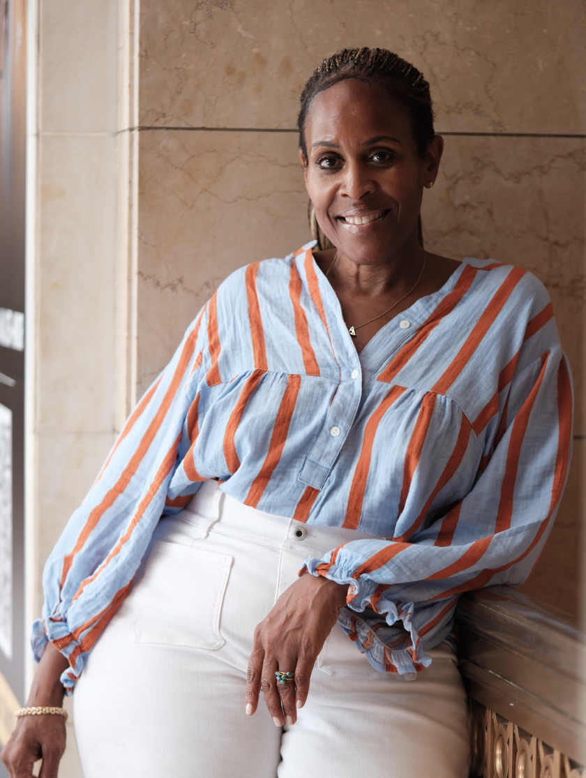 Woman in striped blouse and white pants smiling, leaning against a marble wall indoors.