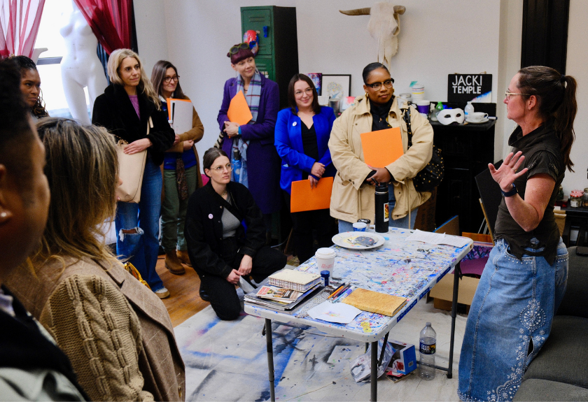 A group of people listens to a woman speak in a colorful art studio