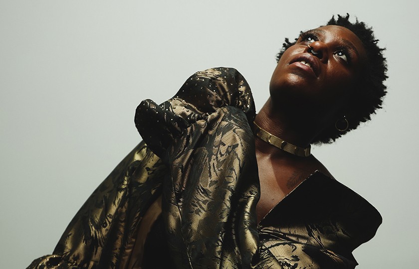 A Black woman with short dark hair, a nose ring, necklace, and gold outfit twists her body while posing dramatically