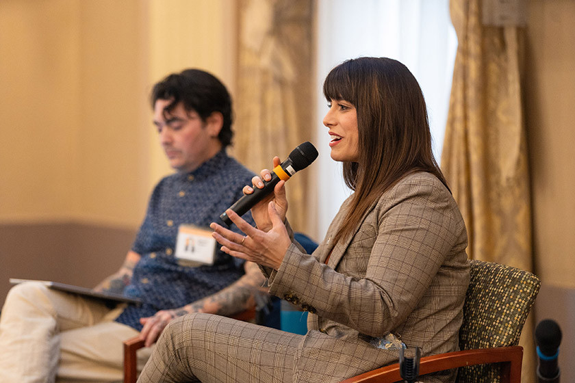 Woman in a suit speaks into a microphone in the foreground beside a man in a button down shirt