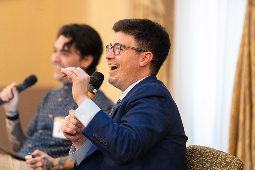 Two men sit and speak into microphones, one laughing while wearing a blue suit.