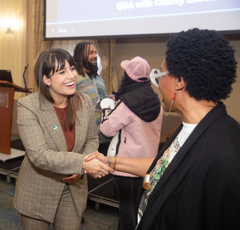 Two women shake hands and smile during an indoor event with others nearby.