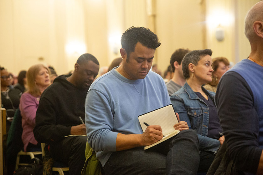A group of people take notes while sitting on chairs in a crowded rom