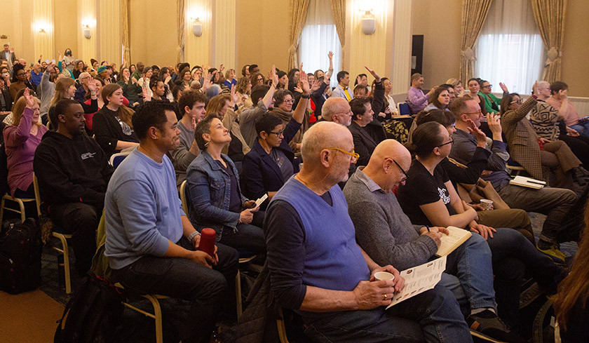 Large group of people seated indoors, some raising hands and listening attentively.