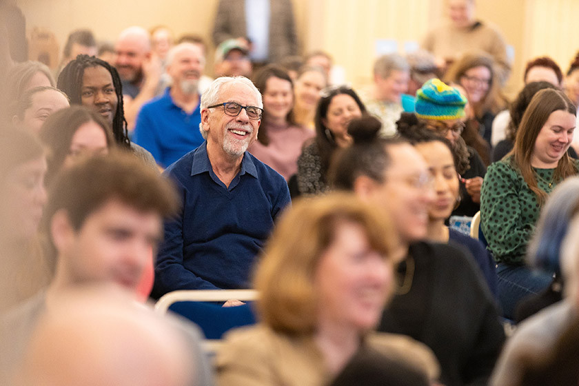 A group of people sit and smile while attending an indoor event or gathering.