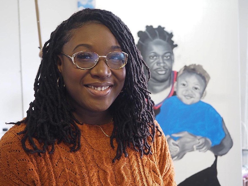 A smiling Black woman with long dark hair, glasses, and an orange sweater. Behind her is artwork of a man holding a young child