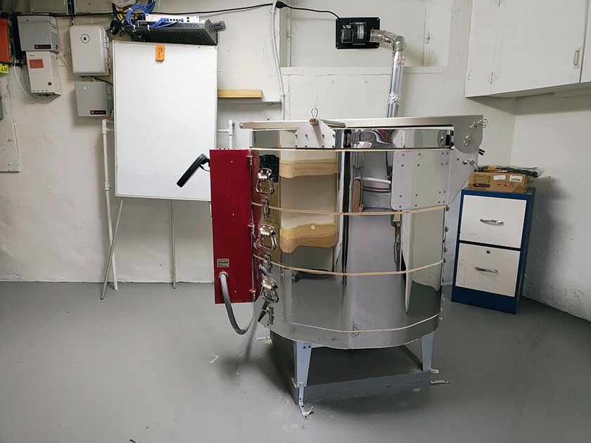 A shiny electric pottery kiln with a red control box stands in a clean, white-walled room with gray floors, next to a whiteboard, cabinets, and some storage shelves.