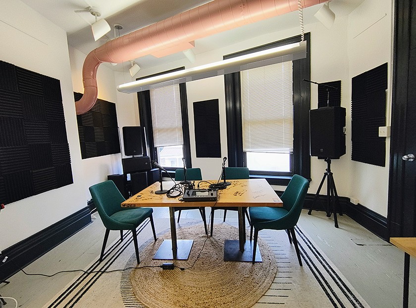A modern podcast studio with four green chairs around a wooden table, microphones, audio equipment, soundproofing panels on the walls, large windows with blinds, and speakers in the corners.