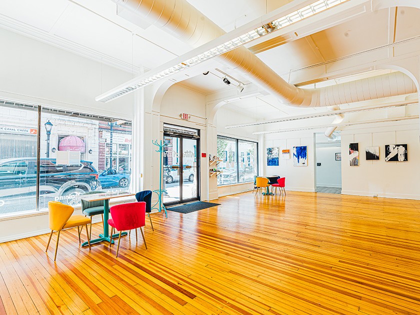 Bright art gallery with large windows, polished wooden floors, colorful chairs around small tables, and contemporary artwork displayed on white walls. Natural light fills the open, airy space.