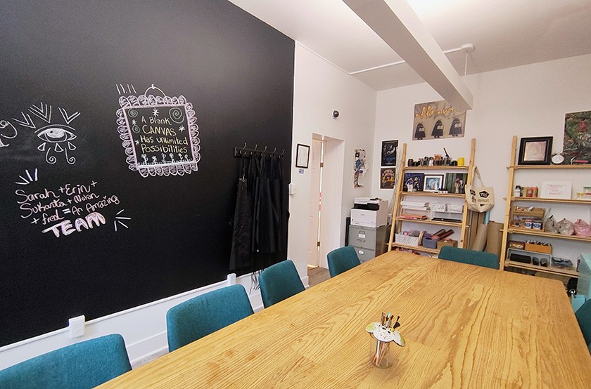 A bright, modern meeting room with a large wooden table, teal chairs, a blackboard wall with chalk drawings, and shelves holding art supplies, books, and decorations. Aprons hang beside the blackboard.