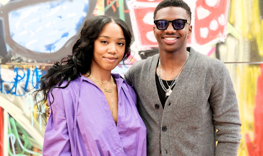 A woman in a loose purple shirt stands next to a man wearing sunglasses, a gray cardigan, and layered necklaces. They are smiling and posing in front of a colorful, graffiti-covered wall.