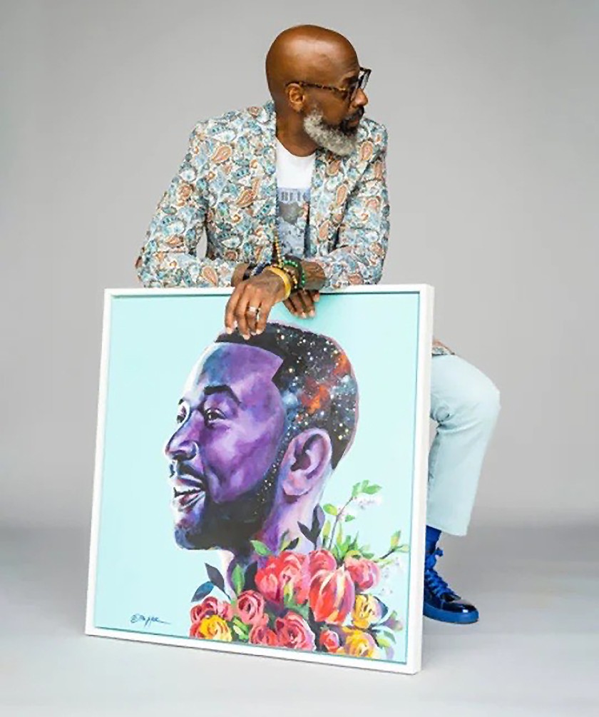 A bald Black bearded man wearing glasses, a white t-shirt, a patterned blazer, light slacks, and blue-and-black shoes. He's sitting down and posed in front of a large framed illustration of a man with flowers
