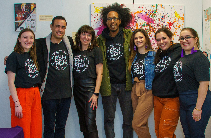 Seven people in matching shirts stand smiling together in front of colorful
