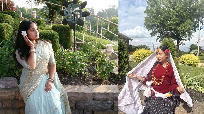 Two women are seated outdoors in traditional attire. The woman on the left wears a pale blue saree and poses with a flower in her hair. The woman on the right wears a maroon top, patterned skirt, and white shawl with jewelry.