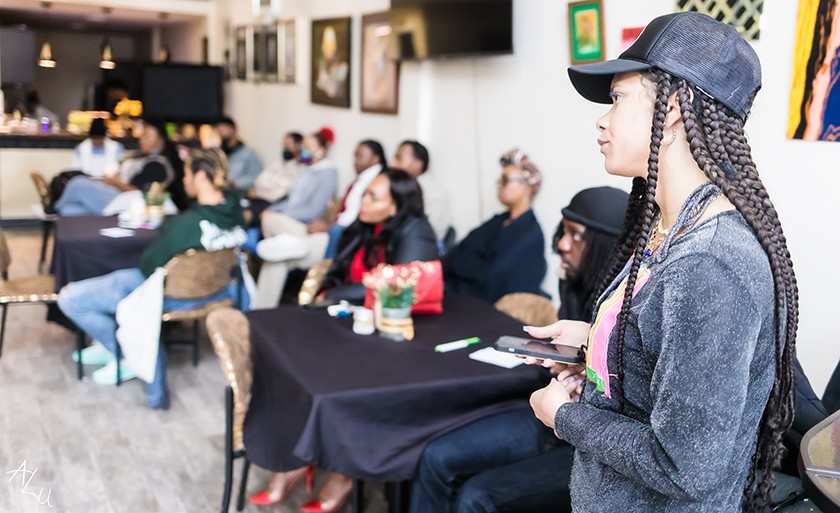 A Black woman with long hair and a baseball cap stands in the foreground in front of a room full of folks watching a panel discussion