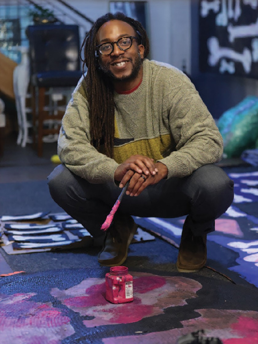 A Black man with long hair, black glasses, jeans, and a green sweater smiles while kneeling down and holding a paint brush, surrounded by paintings on the floor