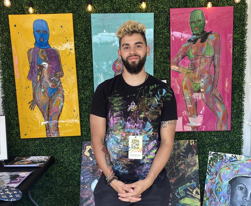 Man with dark beard and bleached hair smiles in front of artwork in a booth