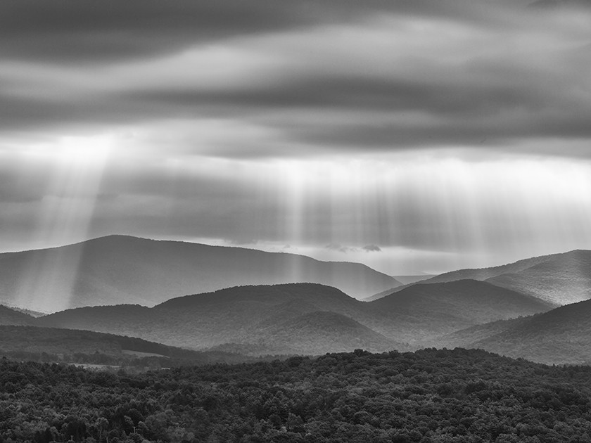 A black-and-white photograph showing beams of light coming down from a cloudy sky and falling behind rows of hilly mountaintops