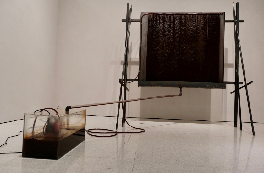 Art installation with a dark liquid circulating from a tank to a large upright rectangular panel.