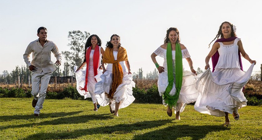 One man in white pants and button-down shirt and four women dressed in loose white dresses and colorful scarves laugh as they run through an outdoors grassy area towards the camera