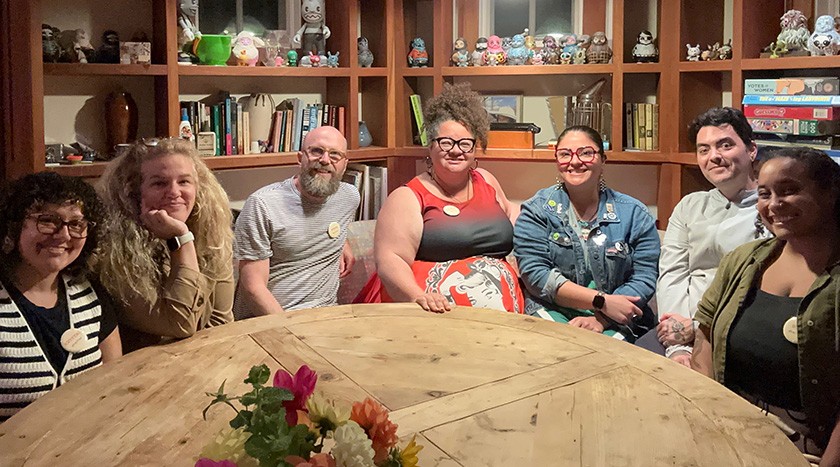 A group of seven people sit around a round wooden table, smiling at the camera. Books and colorful figurines are displayed on shelves behind them. 