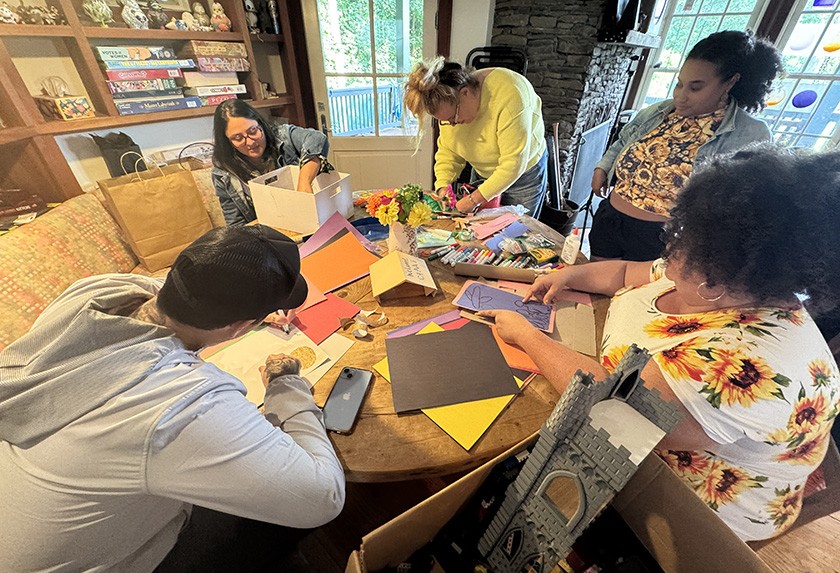 Five people sit around a wooden table covered with colorful paper, markers, and craft supplies, engaging in arts and crafts together in a cozy, well-lit room with shelves and a window in the background.