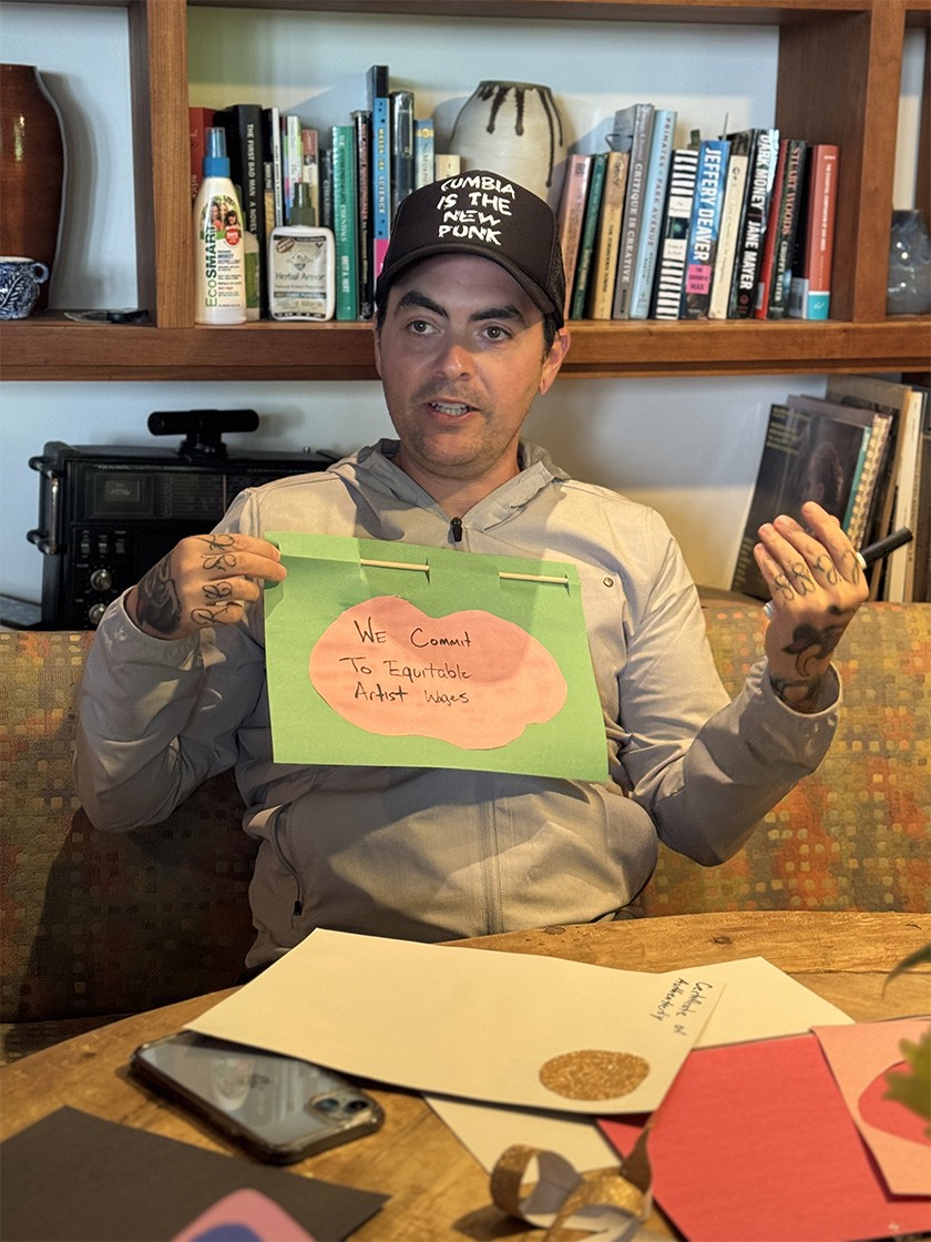 A person holds a handmade sign that reads We commit to equitable artist wages, sitting at a table with craft supplies and bookshelves in the background.