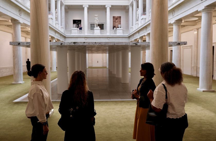 Four people stand talking in a grand museum hall with columns and artwork on display.